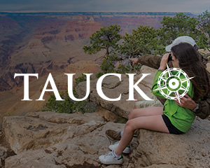 Two people embrace as they sit on a cliff looking over a grand valley, with the Tauck logo superimposed over them.